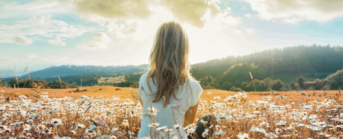 A woman sitting in a field of white flowers, looking out towards distant hills beneath a bright, cloudy sky.